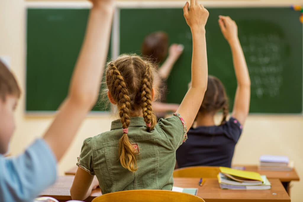 Young children facing the teacher with arms raised to answer a question