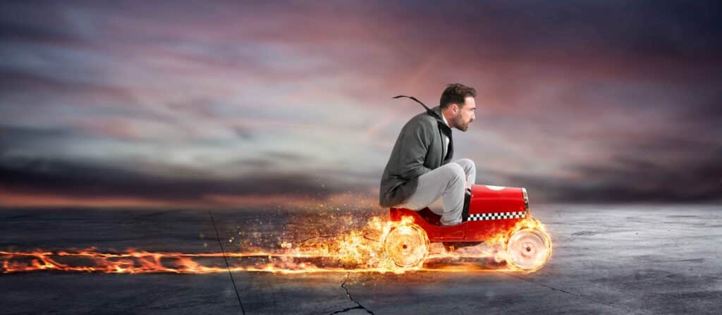 Younger man driving a children's toy car with flames coming off the wheels.