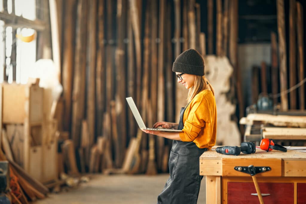 Young female carpenter in woodworking shop looking at laptop
